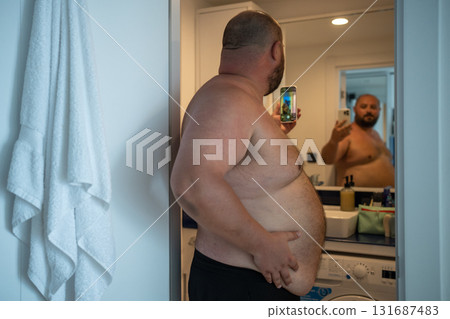 Overweight man with naked torso taking selfie on smartphone looking at mirror in bathroom at home. Overweight man with naked torso taking selfie on smartphone looking at mirror in bathroom at home. 131687483