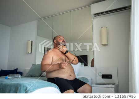 Overweight bearded man in shorts in an air-conditioned room escaping from heat in hotel. 131687486