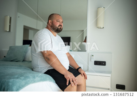 Fat guy sitting on bed with disgruntled face. Guy woke up from heat stuffiness high temperature Fat guy sitting on bed with disgruntled face. Guy woke up from heat stuffiness high temperature 131687512