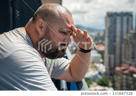 Man covers with hand from sun look into distance from balcony. Fat bald bearded guy hiding with palm 131687526