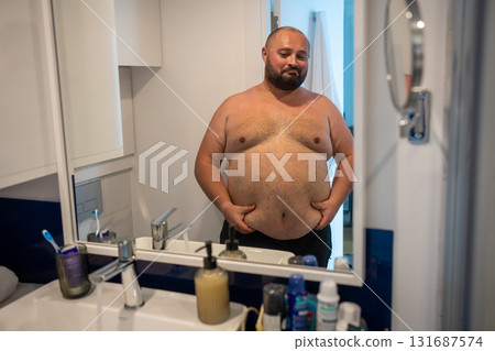 Fat man looking at mirror smiling touching big abdomen in bathroom at home. Fat man looking at mirror smiling touching big abdomen in bathroom at home. 131687574