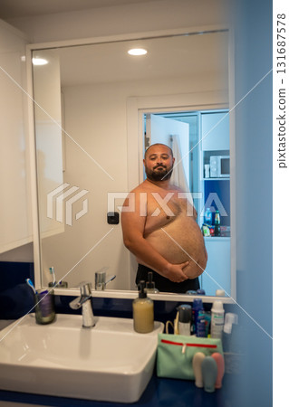 Overweight smiling man with naked torso suffering from extra weight looks at mirror touches abdomen. 131687578