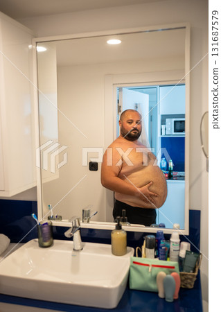 Overweight smiling man with naked torso suffering from extra weight looks at mirror touches abdomen. Overweight smiling man with naked torso suffering from extra weight looks at mirror touches abdomen. 131687579