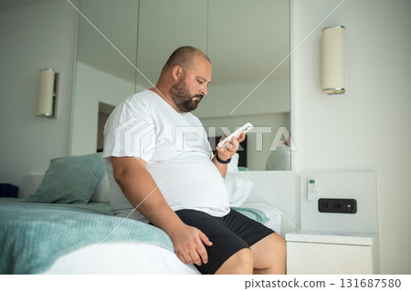 Overweight guy hanging in smartphone on resort sitting in bed in hotel room on vacation. 131687580