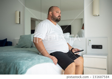 Lazy overweight man sits on hotel bed with phone, gaze thoughtfully at one point. Loneliness, fear Lazy overweight man sits on hotel bed with phone, gaze thoughtfully at one point. Loneliness, fear 131687584