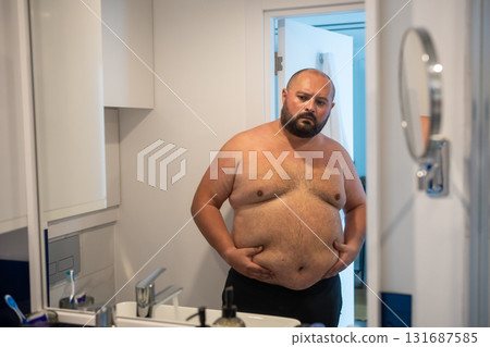 Fat obese guy touching stomach in front mirror dissatisfied with overweight concerned about figure 131687585