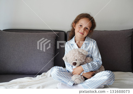 A little girl in blue and white striped pajamas sits on a bed holding a fluffy teddy bear and smiling softly 131688073