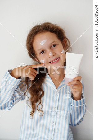 A cheerful little girl in striped blue pajamas applies cream to her face while holding a white cosmetic tube 131688074