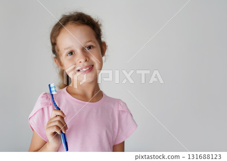 A smiling little girl in a pink t-shirt holds a blue toothbrush, promoting healthy dental hygiene habits 131688123