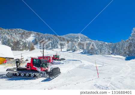 A red snow groomer parked beside a ski slope and a mountain covered in frost (Yokoteyama, Shiga Kogen, Nagano Prefecture) 131688401