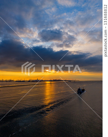 Aerial sunset view of industrial seascape ship leaves wake across golden water as cranes and port structures silhouette against dramatic clouds and warm horizon light. 131688812