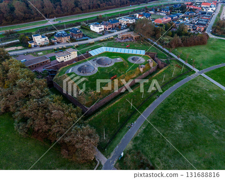 Aerial view of Fort bij Veldhuis grass covered bunkers within brick walls, adjacent to glass roofed museum, surrounded by fields, homes, and highway, blending heritage with modern infrastructure. 131688816