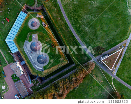 Aerial view of Fort bij Veldhuis grass covered bunkers within brick walls, adjacent to glass roofed museum, surrounded by fields, homes, and highway, blending heritage with modern infrastructure. Aerial view of Fort bij Veldhuis grass covered bunkers within brick walls, adjacent to glass roofed museum, surrounded by fields, homes, and highway, blending heritage with modern infrastructure. 131688823
