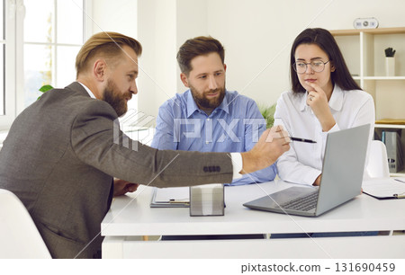 Real estate agent sitting at desk, talking to young couple and showing them something on laptop 131690459