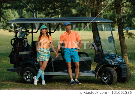 Golfing couple enjoying a sunny afternoon on the course while standing by their golf cart in a scenic park setting 131691586
