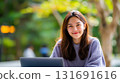 Smiling Asian woman with long brown hair outdoors. A young Asian woman smiles warmly, sitting outside with a laptop. The background has soft green foliage, creating a natural, inviting atmosphere. 131691616