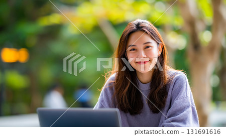 Smiling Asian woman with long brown hair outdoors. A young Asian woman smiles warmly, sitting outside with a laptop. The background has soft green foliage, creating a natural, inviting atmosphere. 131691616