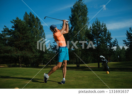 Man swings golf club on sunny day at a lush green course surrounded by pine trees, enjoying the sport with a calm blue sky above Man swings golf club on sunny day at a lush green course surrounded by pine trees, enjoying the sport with a calm blue sky above 131691637
