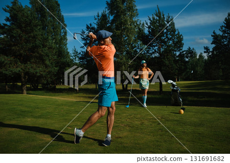 Golfer swings club on a sunny afternoon at a lush course while partner watches nearby with a playful demeanor and a colorful outfit 131691682