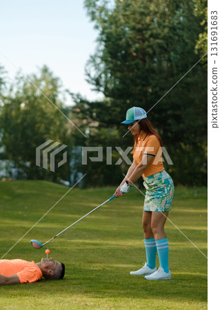 Golfing during a sunny afternoon with a playful twist at a lush green course featuring a humorous moment between a woman and a man on the ground 131691683