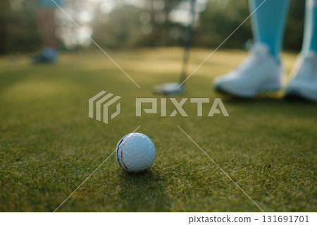 Golf ball on green grass during a sunny afternoon at a golf course with players preparing to take their shots nearby Golf ball on green grass during a sunny afternoon at a golf course with players preparing to take their shots nearby 131691701