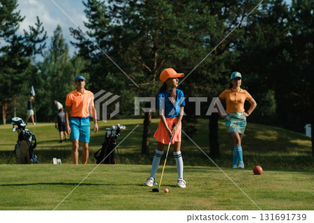 Young girl prepares to putt on a sunny day at the golf course with family watching closely Young girl prepares to putt on a sunny day at the golf course with family watching closely 131691739