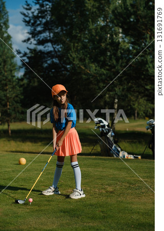 Young girl practices golf swing on a lush green course with trees in the background, dressed in colorful athletic wear during a sunny afternoon Young girl practices golf swing on a lush green course with trees in the background, dressed in colorful athletic wear during a sunny afternoon 131691769