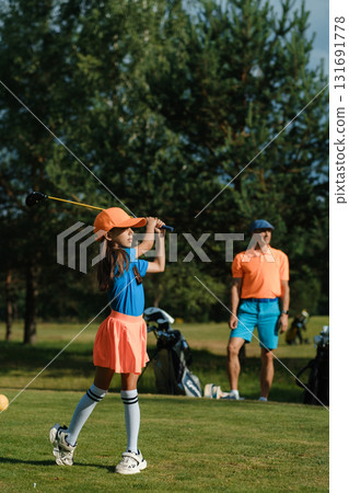 Young girl practices golf swing on a sunny day while an adult observes in matching attire near a golf course 131691778