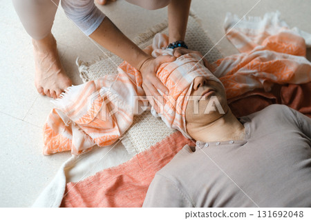 Man lying on the floor while a person ties a cloth around his eyes during a relaxation or meditation practice in a serene indoor setting 131692048