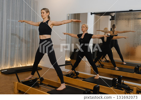 Two women practicing reformer Pilates in a bright, modern studio with mirrors and natural lighting during a morning fitness session Two women practicing reformer Pilates in a bright, modern studio with mirrors and natural lighting during a morning fitness session 131692073