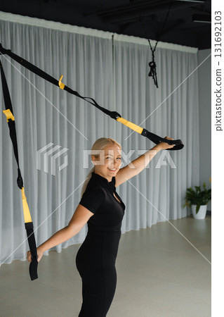 During the day, a woman trains on a suspension in a modern fitness studio, demonstrating strength and posture with yellow straps and a bright smile. 131692103