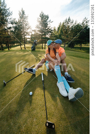 A couple in colorful clothes sits on a golf course and kisses on a sunny day, surrounded by trees and golf equipment A couple in colorful clothes sits on a golf course and kisses on a sunny day, surrounded by trees and golf equipment 131692161