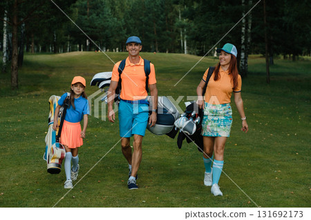 A family in sports clothes and carrying golf clubs walks along the golf course, holding bags in their hands and in bright outfits 131692173
