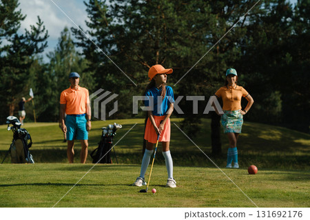 A young girl prepares to hit a ball on a sunny golf course surrounded by her parents, playing a family game of golf on a weekend morning A young girl prepares to hit a ball on a sunny golf course surrounded by her parents, playing a family game of golf on a weekend morning 131692176