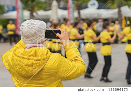 Person Taking Photo of Street Dance. Woman in yellow jacket taking photo of outdoor street performance with smartphone. Concept of autumn, urban lifestyle, hobby, public event, street photography 131694173