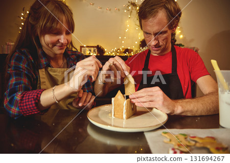 Family Baking Fun: Couple Decorates Christmas Gingerbread House Together During Cozy Holiday Evening Family Baking Fun: Couple Decorates Christmas Gingerbread House Together During Cozy Holiday Evening 131694267