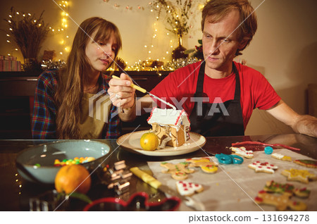 Family Christmas Baking: Couple Decorates Gingerbread House Together in Festive Kitchen with Lights, Cookies, and Christmas Cheer 131694278