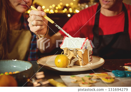 Couple Decorating A Gingerbread House With Icing For Festive Holiday Baking At Home Couple Decorating A Gingerbread House With Icing For Festive Holiday Baking At Home 131694279