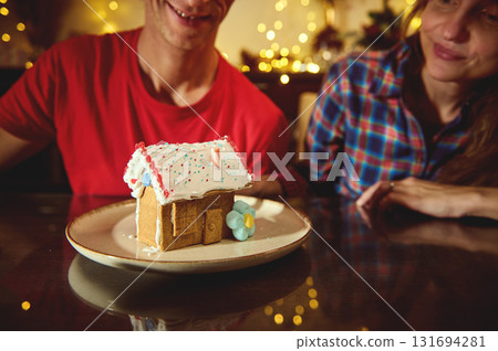 Couple Enjoying Christmas Moment With Gingerbread House Dessert On Plate And Festive Lights In Background Couple Enjoying Christmas Moment With Gingerbread House Dessert On Plate And Festive Lights In Background 131694281