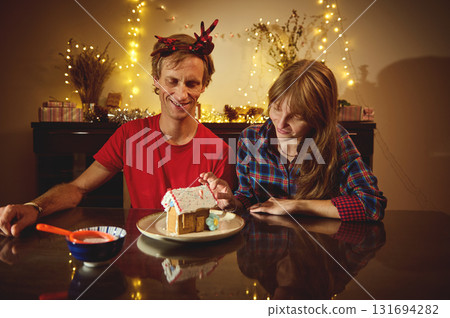 Happy Couple Celebrates Christmas Evening, Decorating Gingerbread House With Warm Lights and Cozy Festive Decor Happy Couple Celebrates Christmas Evening, Decorating Gingerbread House With Warm Lights and Cozy Festive Decor 131694282