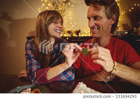 Joyful Couple Sharing Heart-Shaped Cookie by Christmas Lights in a Cozy Home Celebration Joyful Couple Sharing Heart-Shaped Cookie by Christmas Lights in a Cozy Home Celebration 131694293