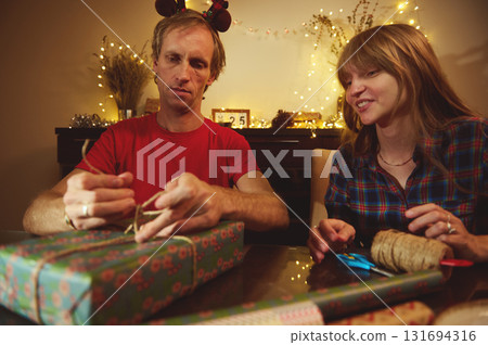 Couple Christmas Gift Wrapping At Cozy Dinner Table With Fairy Lights And Decorations For A Festive Home Moment 131694316