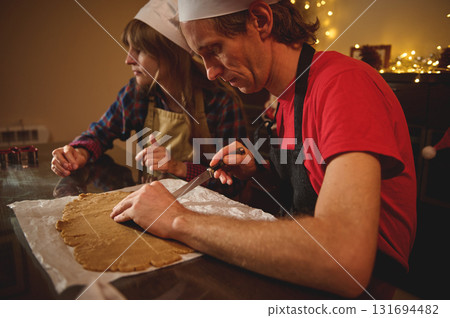 Two Cooks In Aprons Roll Dough For Christmas Treat As Festive Lights Glow In The Background Two Cooks In Aprons Roll Dough For Christmas Treat As Festive Lights Glow In The Background 131694482