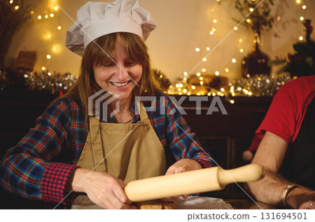 Joyful Baker in Chef Hat and Apron Rolling Dough During Cozy Christmas Kitchen Scene Joyful Baker in Chef Hat and Apron Rolling Dough During Cozy Christmas Kitchen Scene 131694501