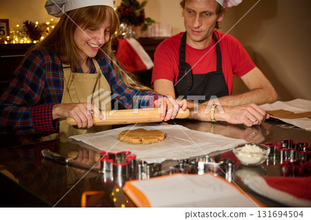 Two People Baking Cookies Together in a Festive Kitchen, Rolling Dough on a Shiny Counter Under Warm Lights, Wearing Chef Hats and Aprons, Christmas Decor in Background 131694504