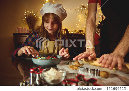 Family Christmas Baking: Child Chef In Apron And Chef Hat Helps Roll Dough And Shape Cookies While An Adult Guides In Cozy Festive Kitchen 131694525