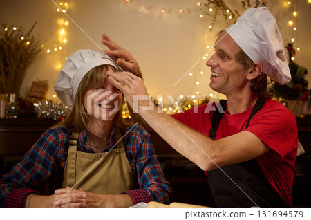Happy Friends In Kitchen: Two Chefs With Hats And Aprons Share Joyful Holiday Moment Under Warm Christmas Lights 131694579