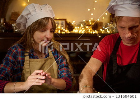 Two Cooks Wearing Chef Hats Prepare Dough At A Festive Kitchen Counter, Sharing Focused Moment Amid Christmas Lights Two Cooks Wearing Chef Hats Prepare Dough At A Festive Kitchen Counter, Sharing Focused Moment Amid Christmas Lights 131694601
