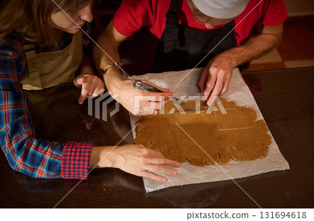 Family And Friends Bake Gingerbread Cookies Together In A Cozy Kitchen, Cutting And Shaping Dough On Parchment For Festive Holiday Treats 131694618