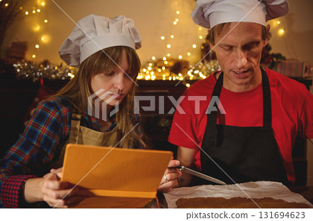Two Home Bakers in Chef Hats Review a Recipe on Tablet While Preparing a Dessert in a Cozy, Festive Kitchen 131694623